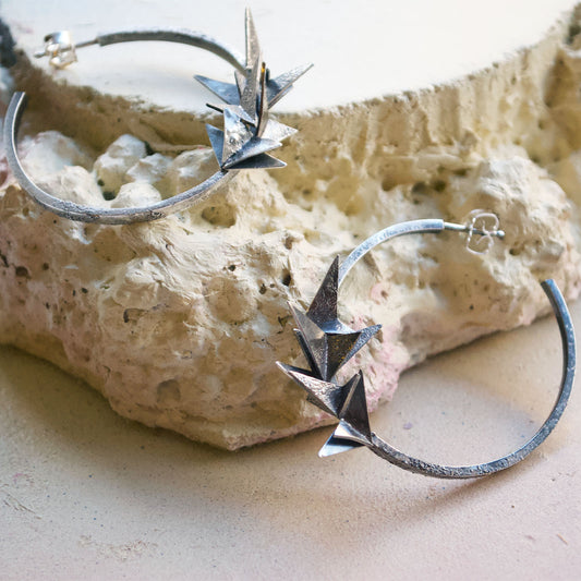 A pair of hoop earrings covered in little silver origami folded planes. The hoops have a rough, rustic, raw texture and are oxidised to look black. Handmade in recycled silver in Copenhagen by Scottish artist Caroline Cloughley.
