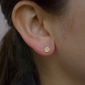 An ear wearing a beautiful small stud earring. The earring is a circle with a moonscape texture resembling a full moon. The texture is rough rustic and organic. Handmade in recycled sterling silver plated with 24ct gold and Handmade in Copenhagen by Scottish designer Caroline Cloughley. The photograph appears to be taken on  a cloudy day but the jewellery appears warm and bright. 